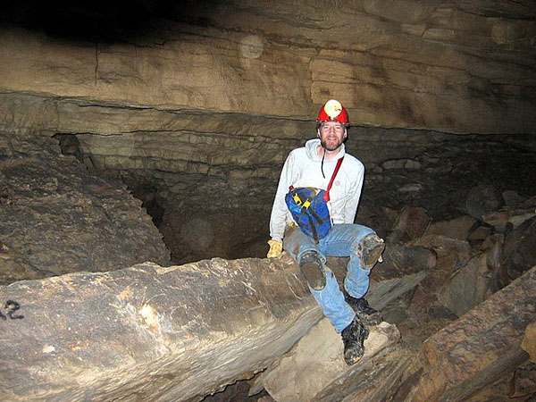 Photo of the author seated on a large rock formation within a cave
							room